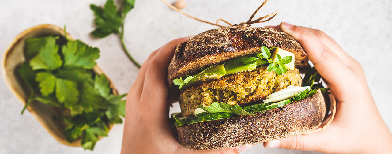 Vegan sandwich with chickpea patty, avocado, cucumber and greens in hands