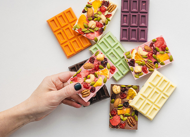 Close up woman's hand holding chocolate bar with inclusions and colorful bars on table