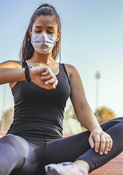 Sporty young woman checking her pulse at the stadium, wearing medical protective mask outdoors
